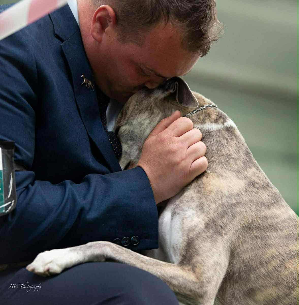 A man gently holding a whippet. The whippet has her head nestled in his chest
